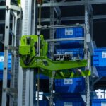Zielone ramię robota przenoszące niebieskie pojemniki w automatycznym magazynie